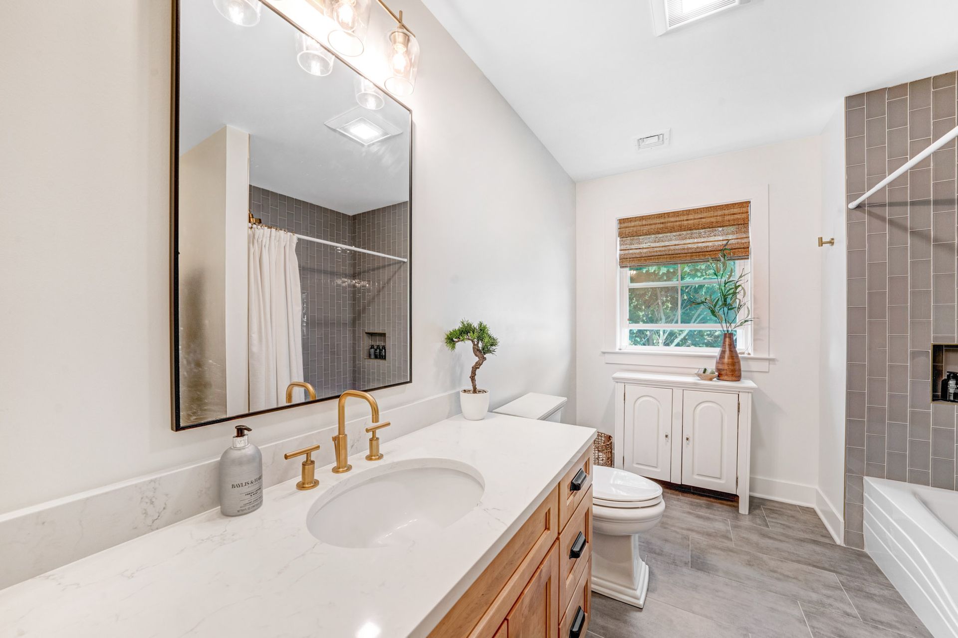 Bathroom with white countertop, gold fixtures, large mirror, shower, and window with bamboo shade.