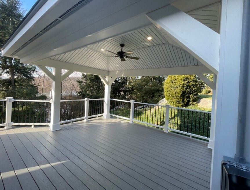 Covered outdoor deck with grey flooring, white beams, ceiling fan, and black railing.
