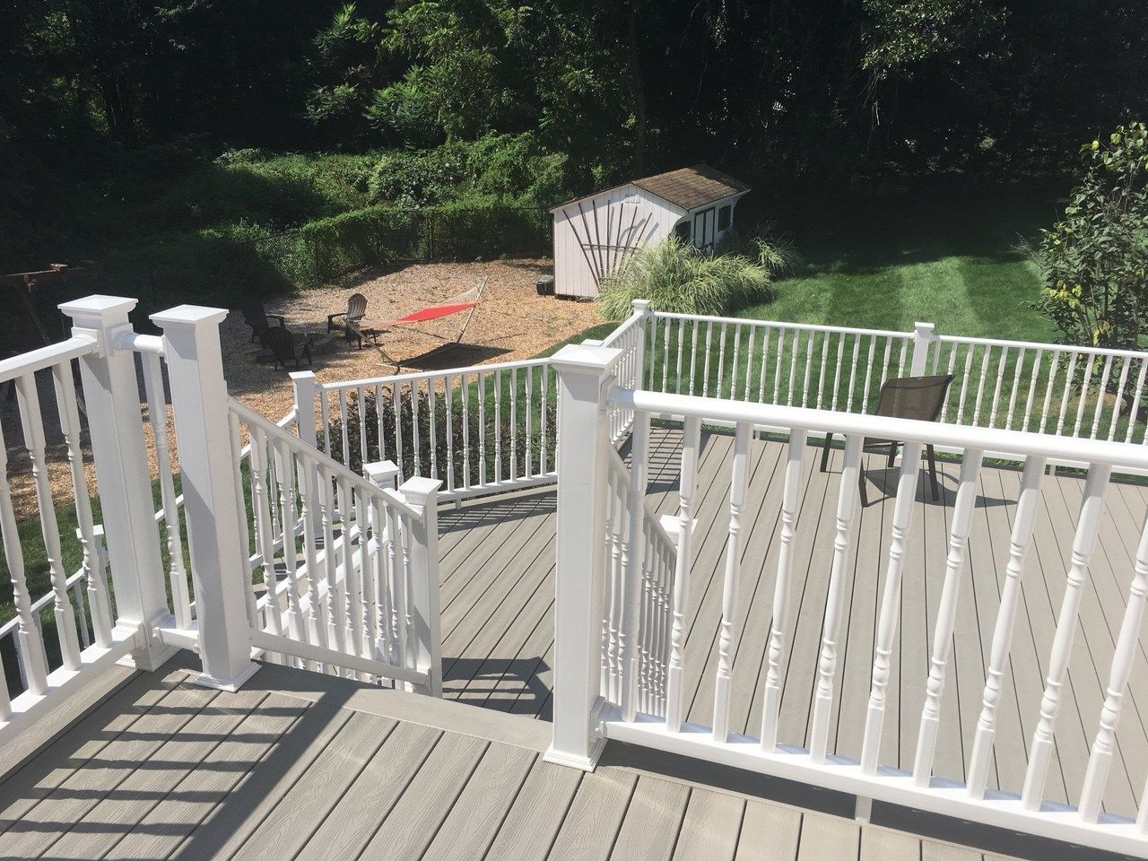 White deck with railings overlooks a backyard with shed, trees, and mowed lawn on a sunny day.