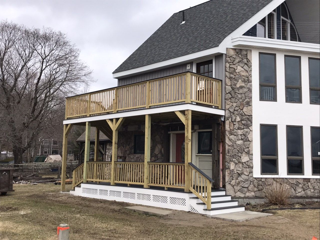 Two-story wooden deck with railing attached to a stone and white house; steps lead to front door.