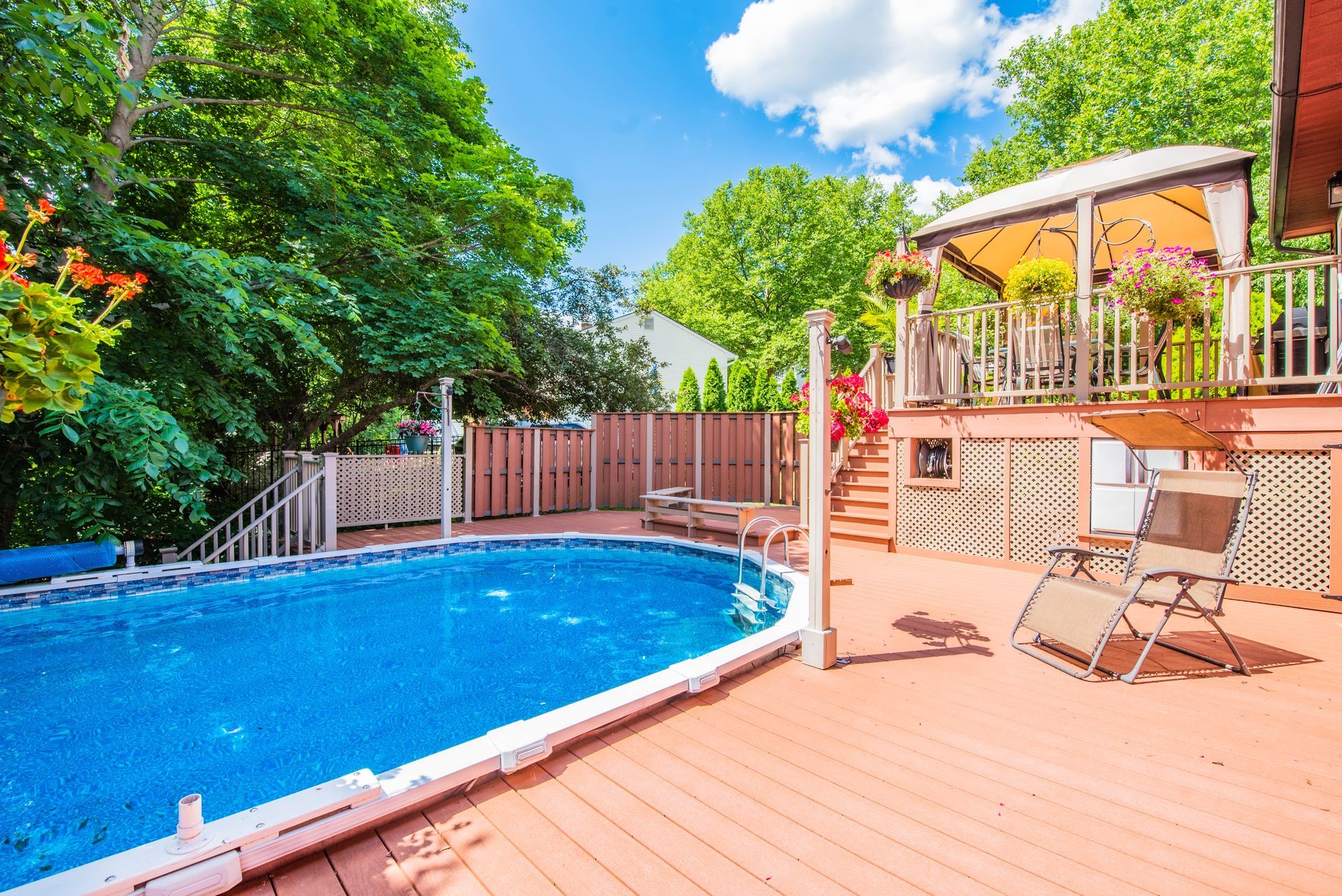 Backyard with above-ground pool, wooden deck, gazebo, and lush greenery under a blue sky.