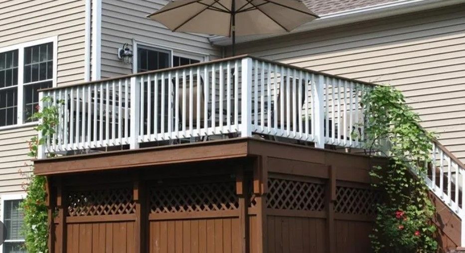 Brown deck with white railing and lattice, beige umbrella, beside a tan house with windows.