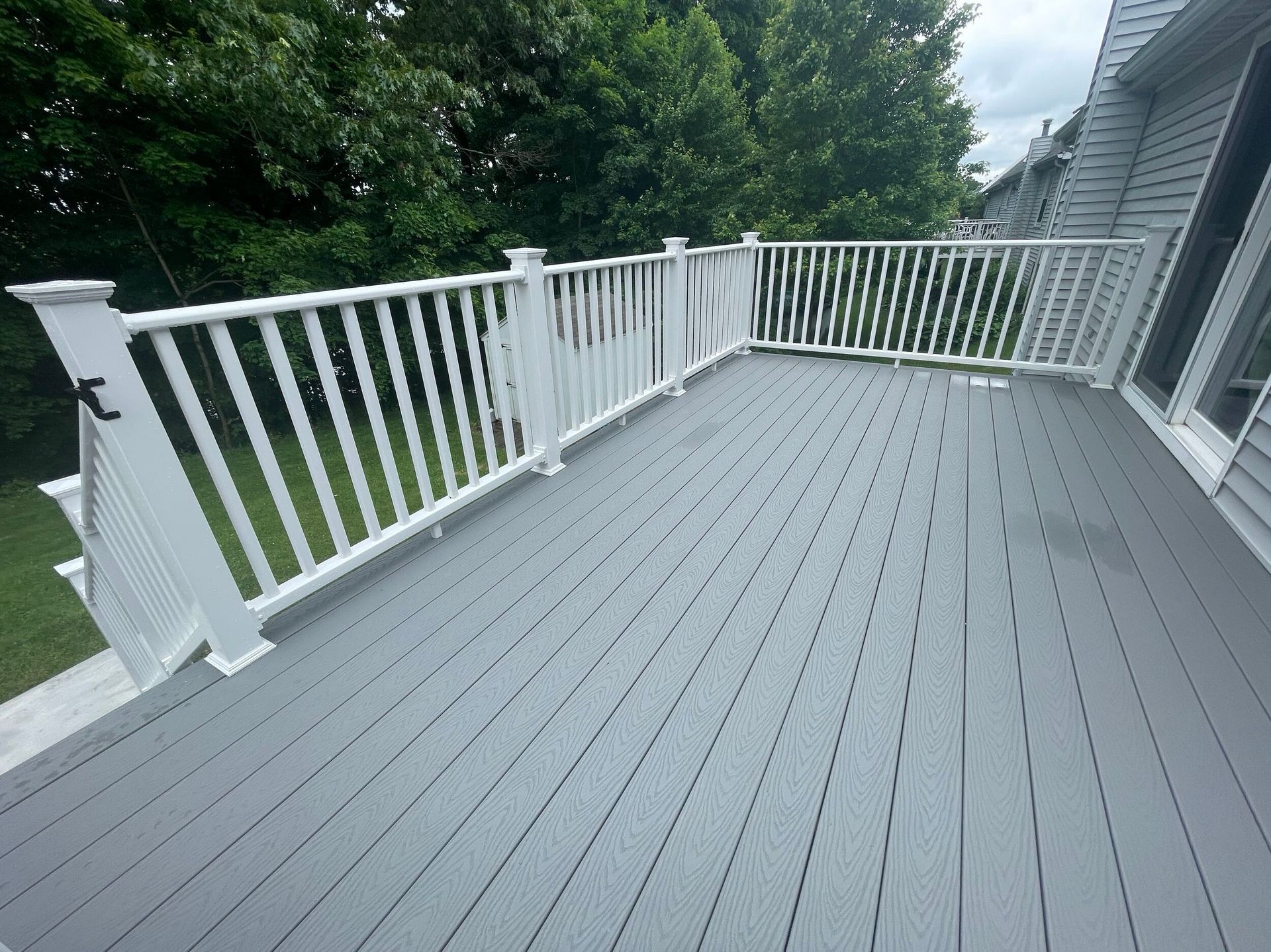 Gray composite deck with white railing, next to a house, surrounded by trees.