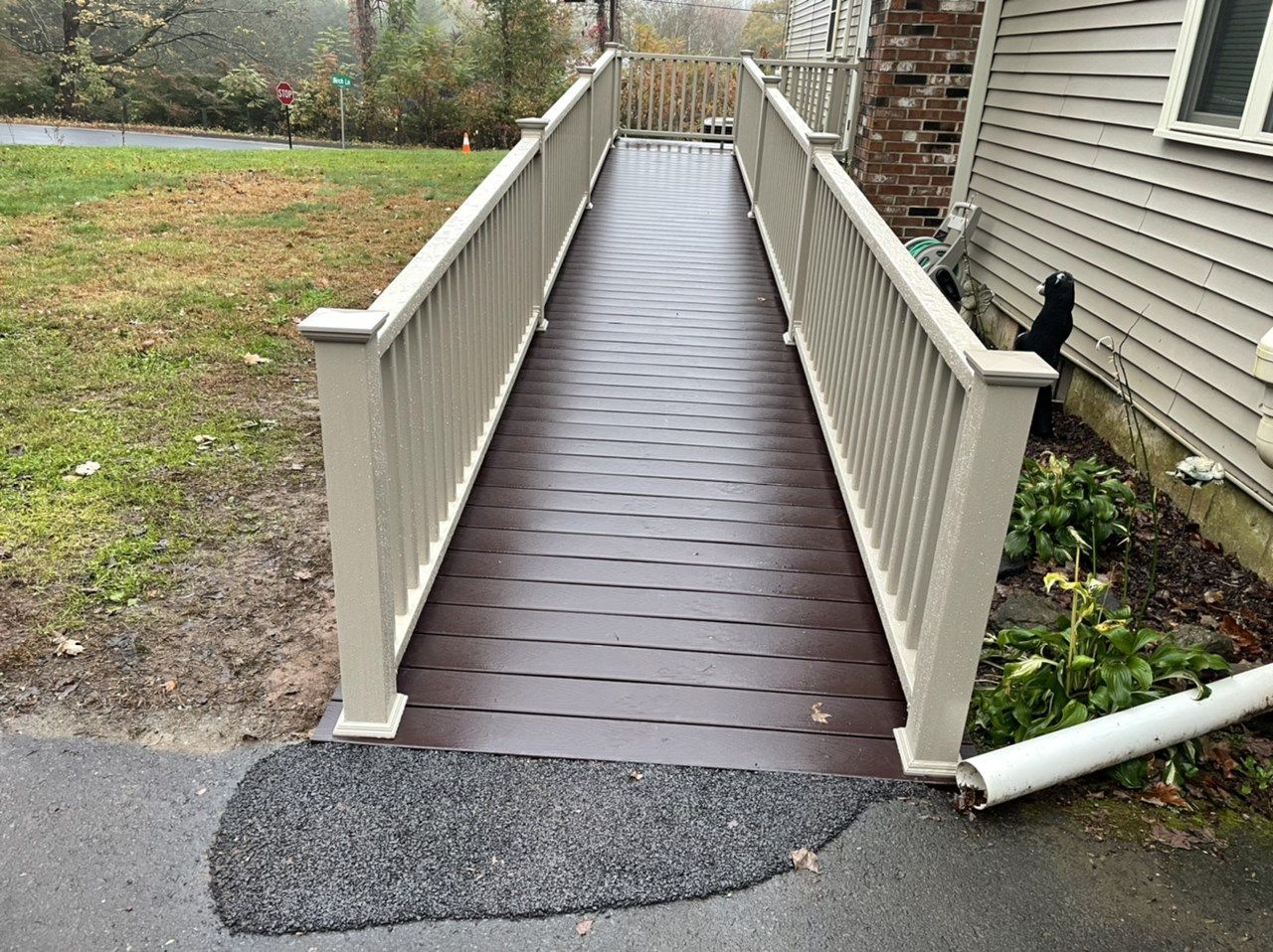 Exterior wheelchair ramp with railing leading to a house entrance. Brown ramp, beige railing, asphalt driveway.