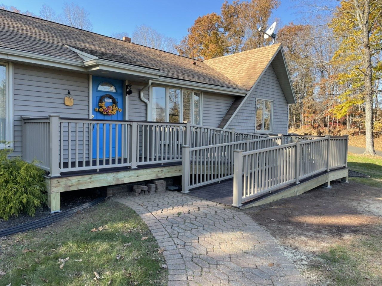 Ramp leading to a light gray house with a blue door and a brick pathway. Autumn foliage surrounds the house.