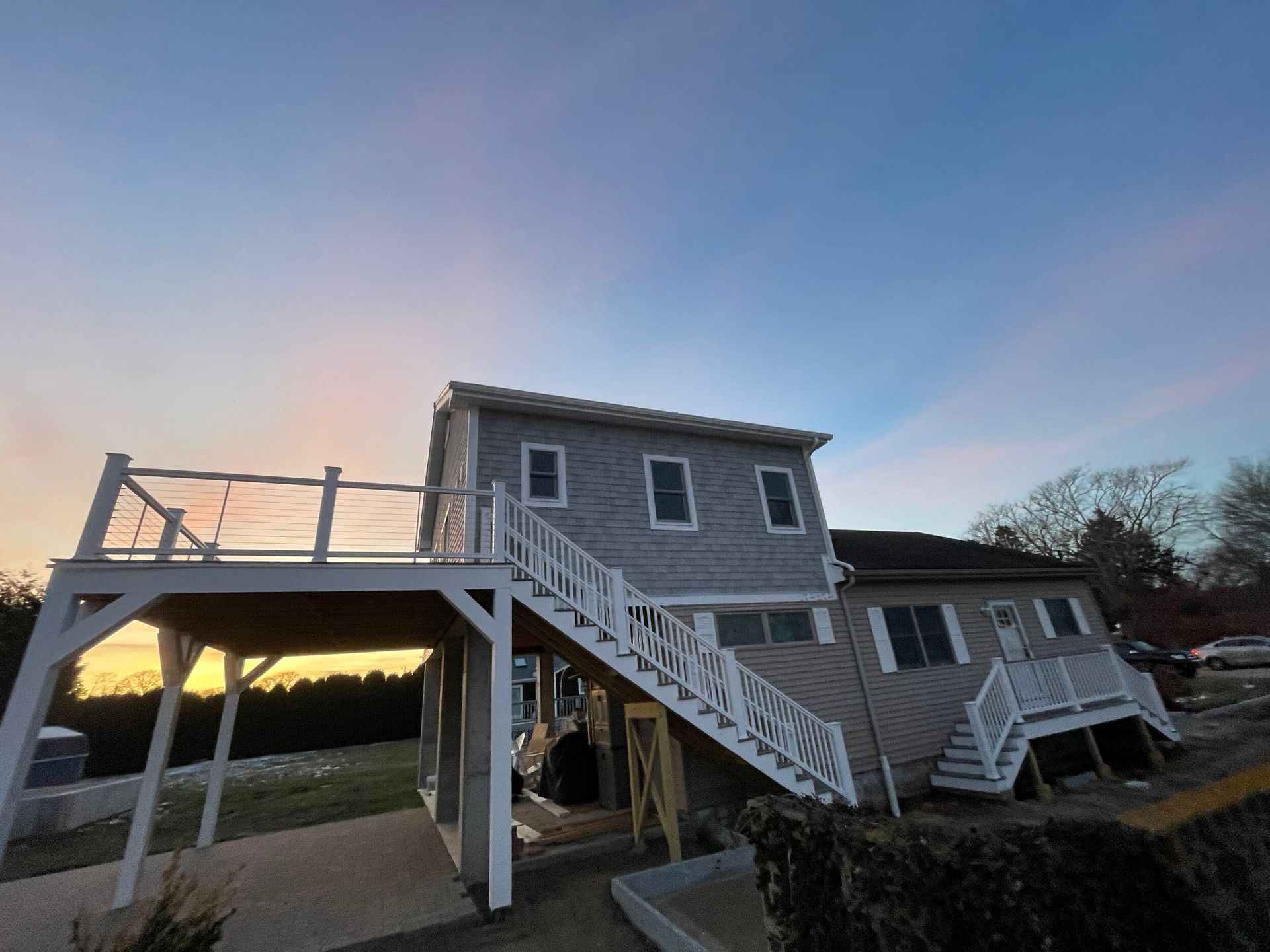 Two-story gray house with deck and stairs against a colorful sunset sky.