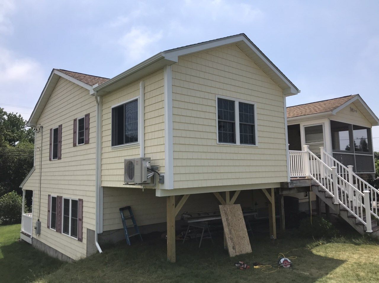 Yellow house on stilts with a deck, next to a smaller screened-in porch, under a blue sky.