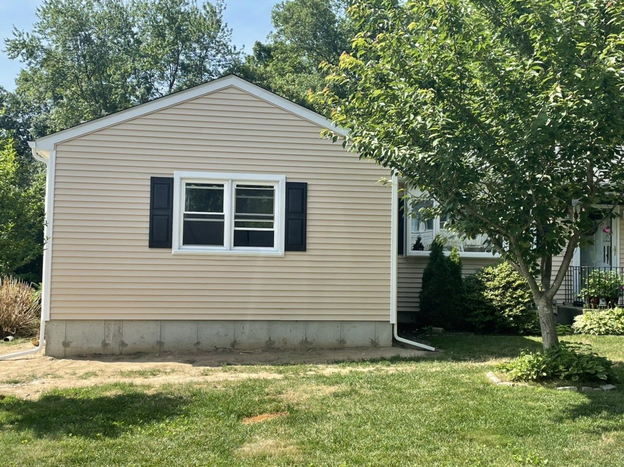 Tan house with a window, black shutters, and a small tree in front. Green grass and blue sky.