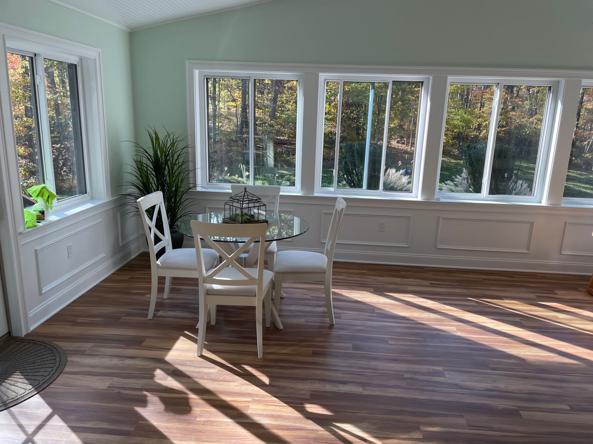 Sunroom with table and chairs. Large windows overlook trees. Brown wood-look floor, green walls, and white trim.