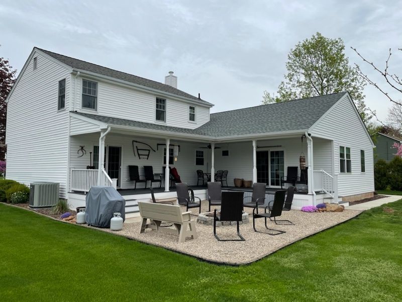 White house with a large backyard patio. Gravel fire pit area with seating. Green grass. Overcast sky.