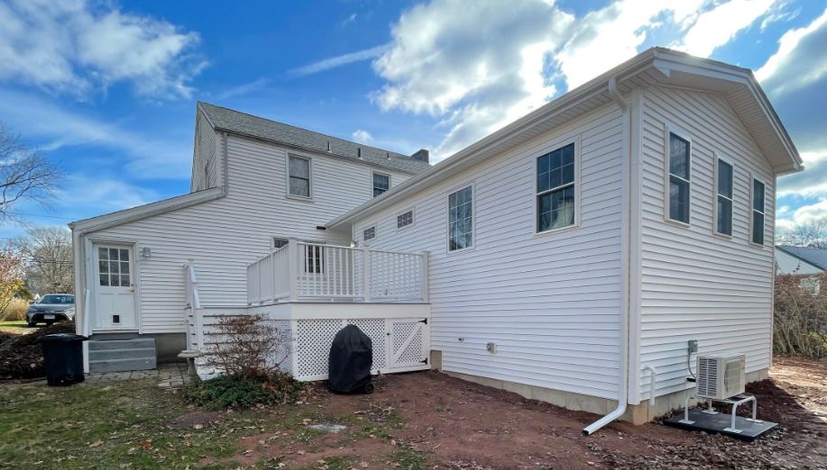 White house with deck, siding, windows, and an air conditioning unit outside, under a blue sky.