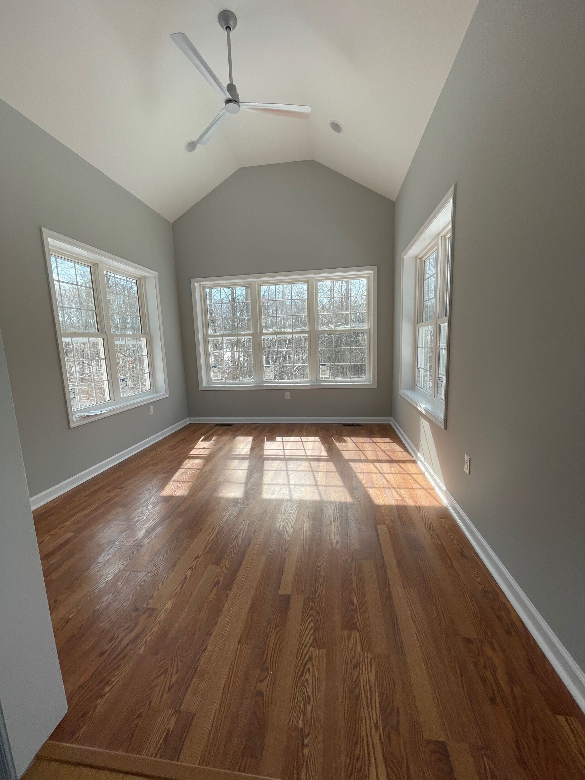 Empty room with hardwood floors, many windows, and a vaulted ceiling. Gray walls and a ceiling fan.
