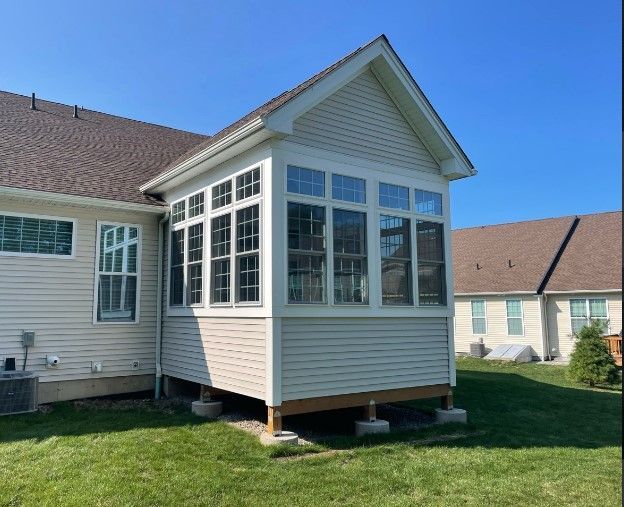A sunroom addition with multiple windows, connected to a beige house with a brown roof, on a grassy lawn under a blue sky.