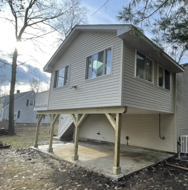 Tan house extension with a deck, supported by wooden beams, over a concrete patio.