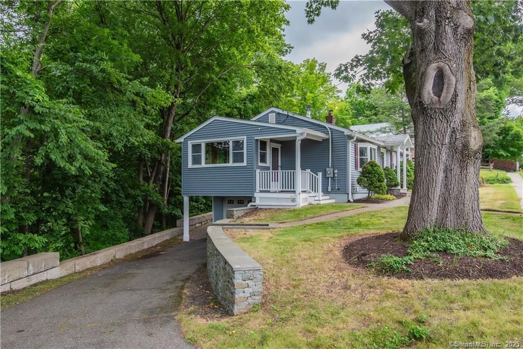 Blue house with white trim, elevated, beside a large tree on a grassy hill; driveway.