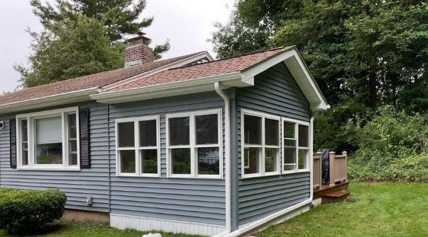 Blue-sided house with a sunroom, white trim, and a brown roof. Green foliage in the background.