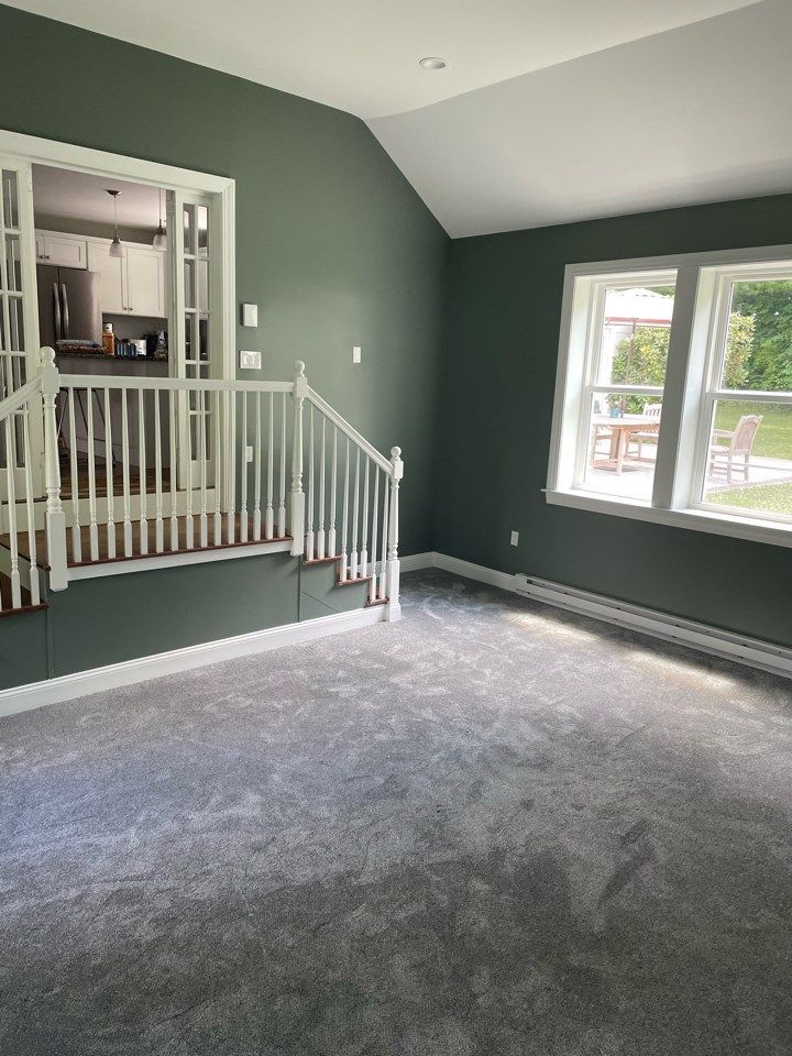 Empty living room with green walls, gray carpet, and white trim. Features stairs and windows.