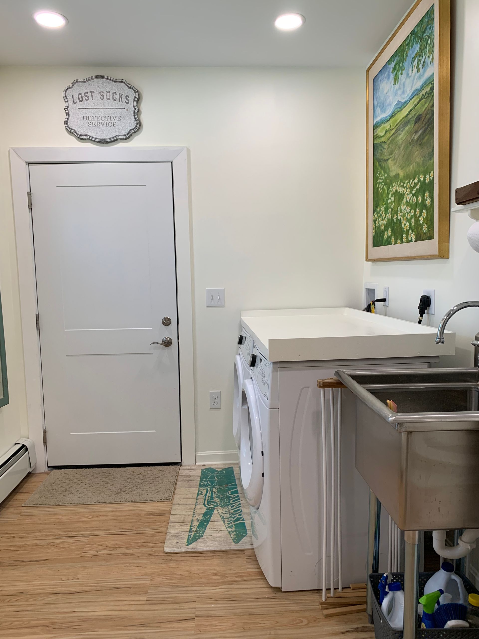 Laundry room with washer, dryer, sink, door, and framed artwork. Light wood-look flooring and cream-colored walls.