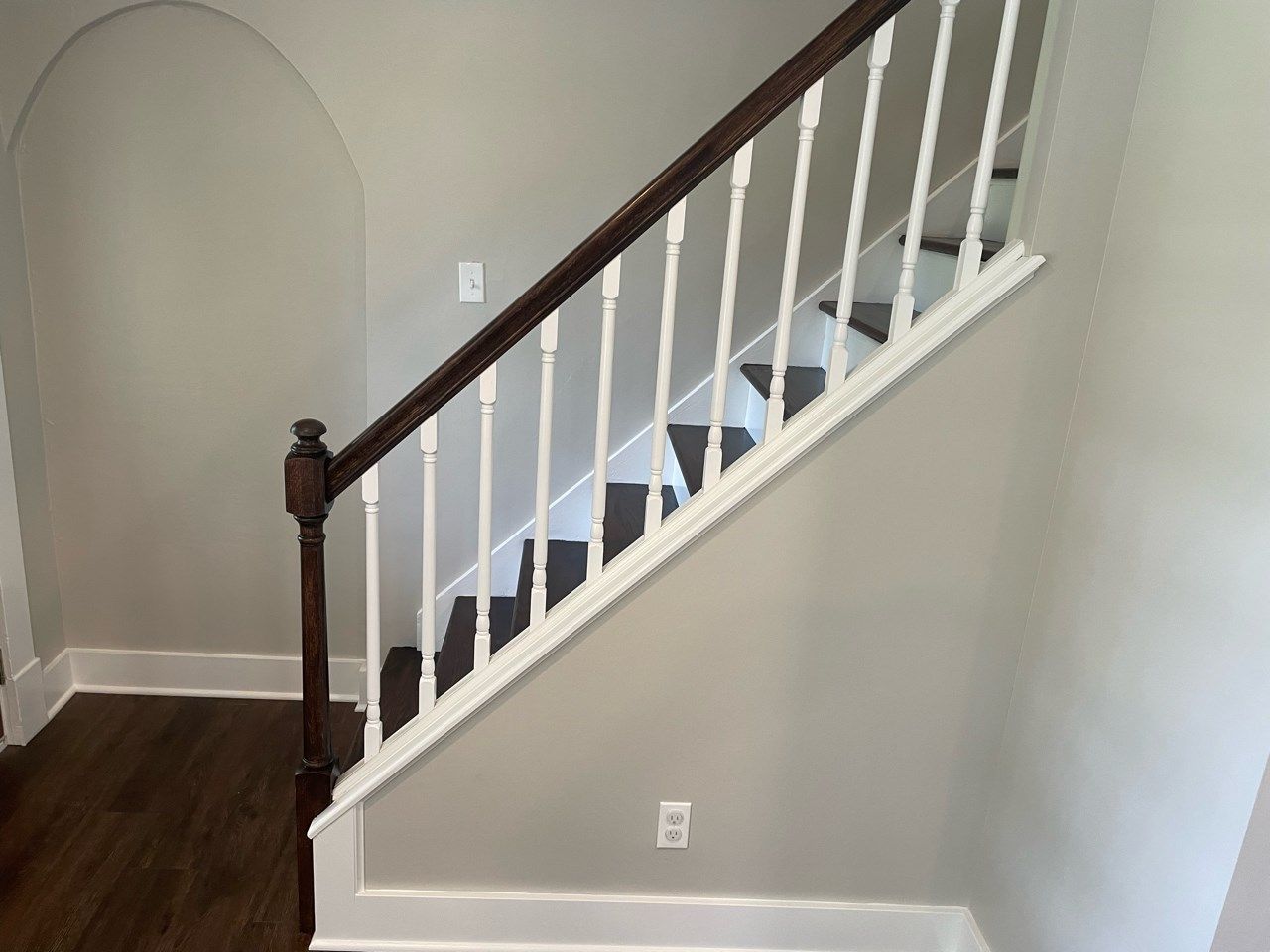 Staircase with white balusters, dark wood handrail, against light gray wall. Dark wood flooring.