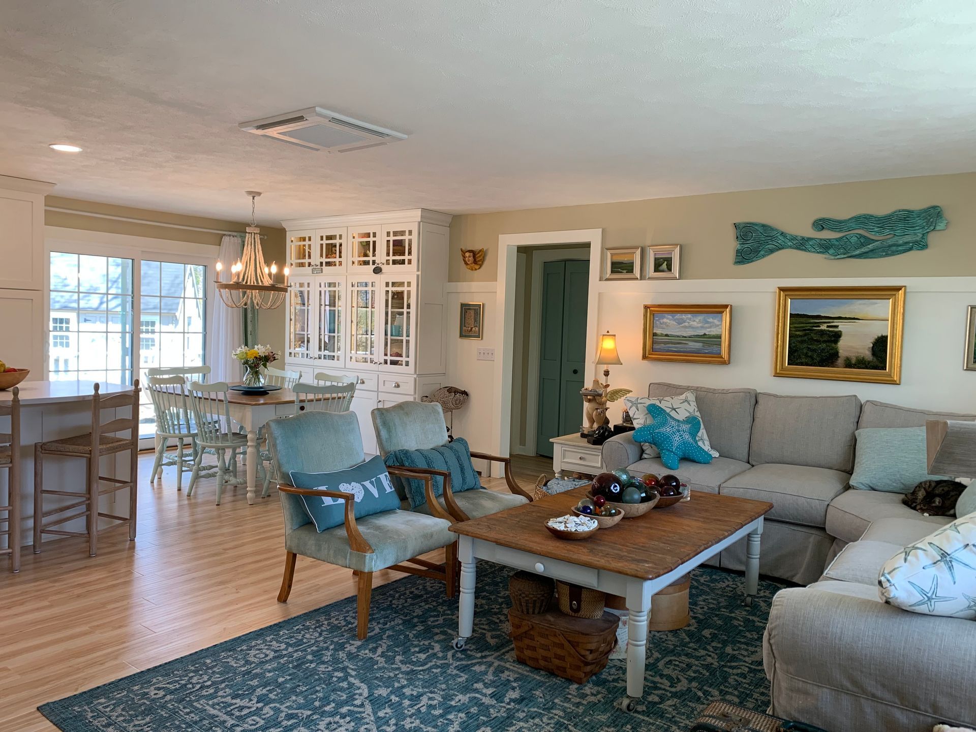 Living room with blue accents, open to dining area and kitchen.  Neutral walls, hardwood floors,  light-colored furniture.