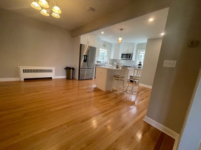 Bright, open-concept living space with hardwood floors. Kitchen visible with white cabinetry and island.