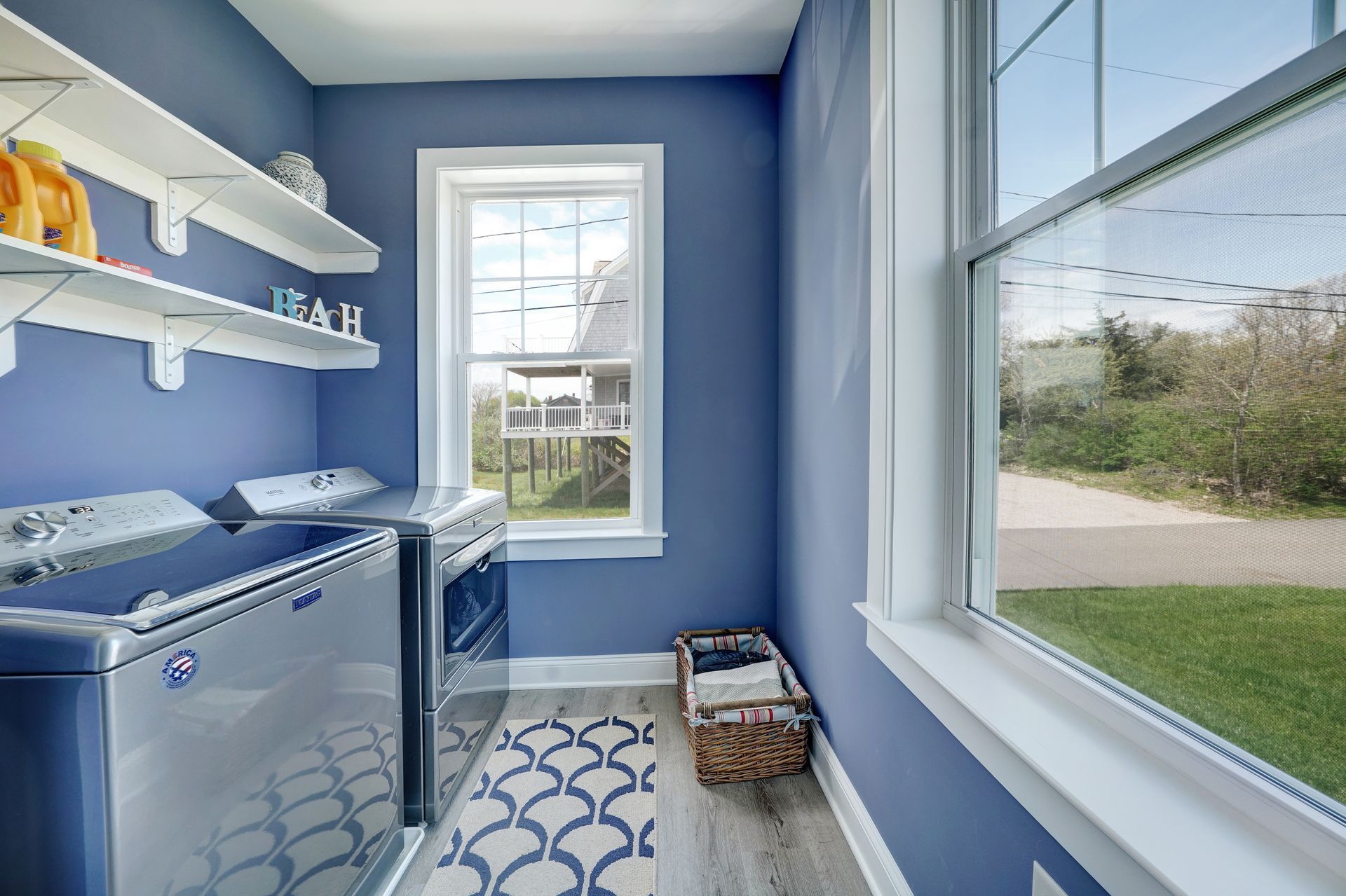 Blue laundry room with washing machine, dryer, shelves, window, and patterned rug.