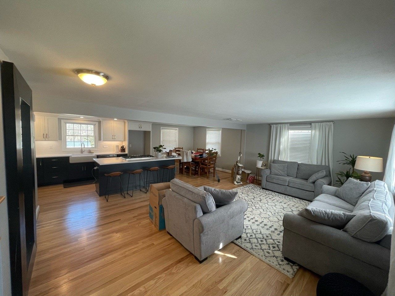 Open-concept kitchen and living room with gray couches, wooden floors, and a kitchen island with bar stools.