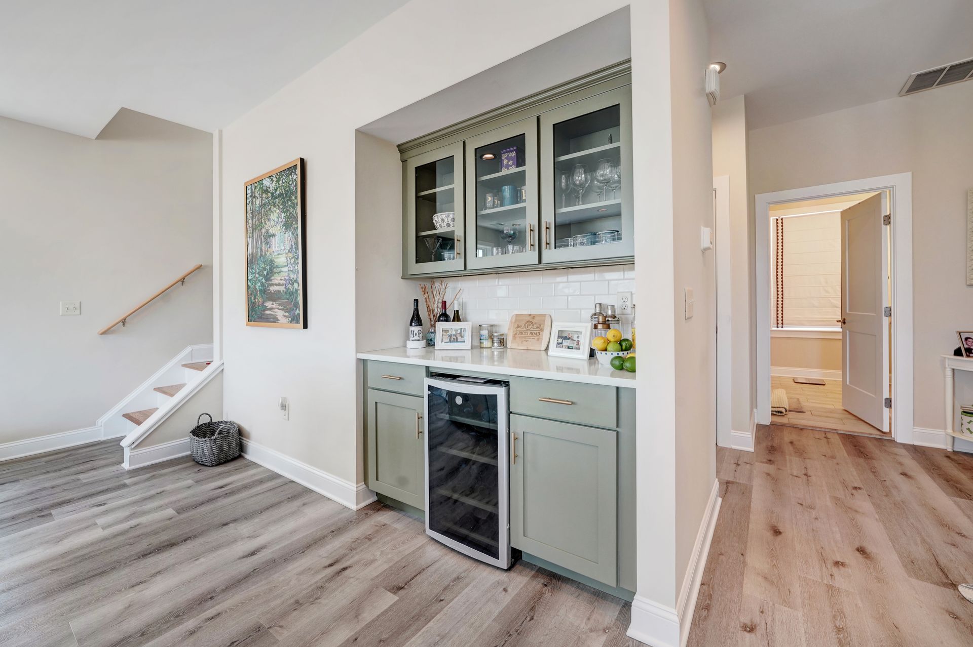 Built-in home bar with sage green cabinets, wine cooler, and glass-front upper cabinets, near a staircase and a bathroom doorway.