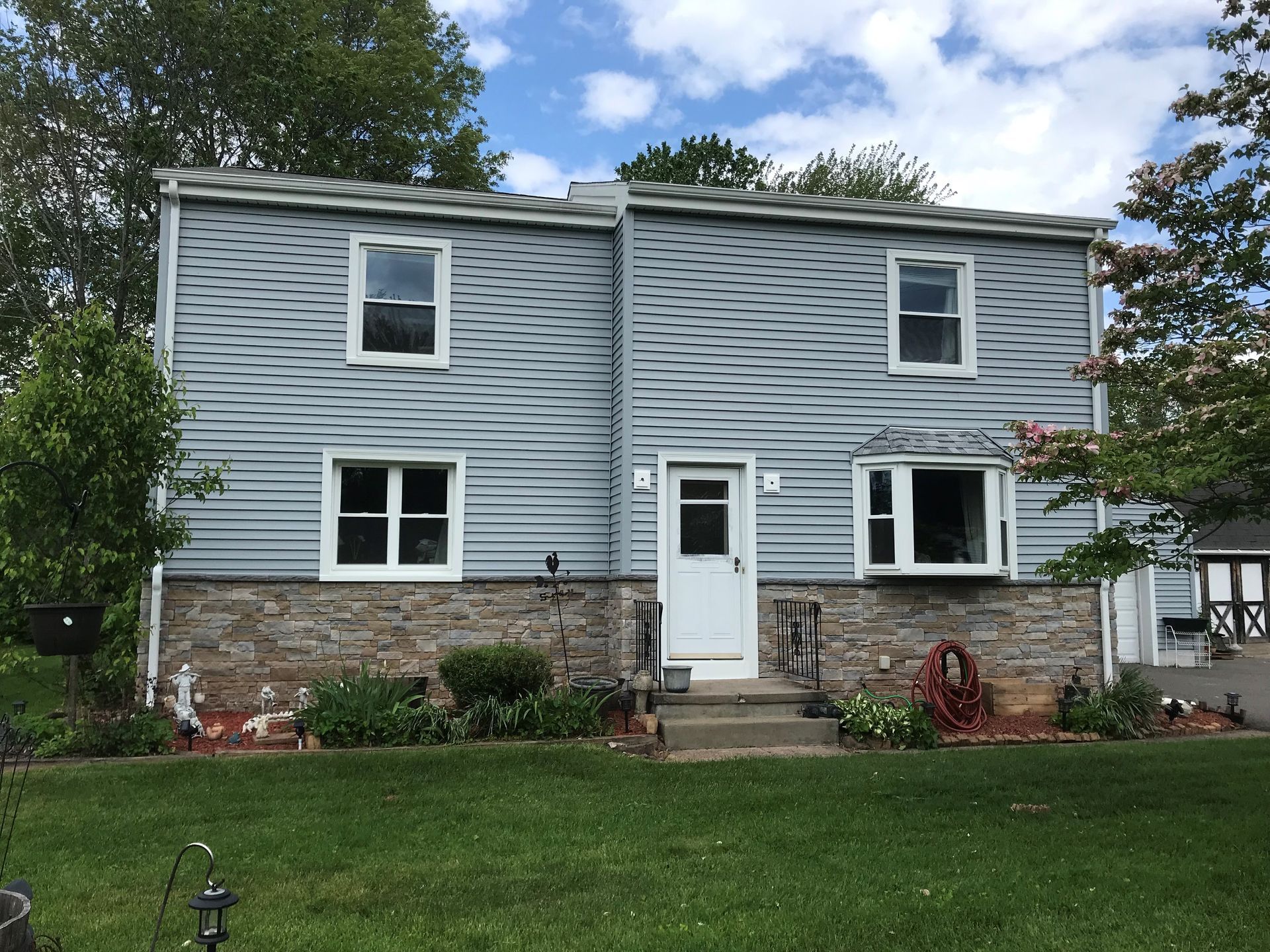 Two-story house with blue siding, stone foundation, and white trim. A front door leads to a small yard.