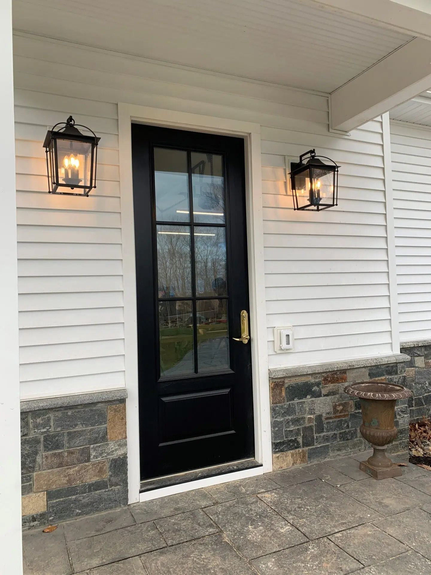 Black front door with glass panels, flanked by two wall-mounted lanterns, on a white house with stone accents.