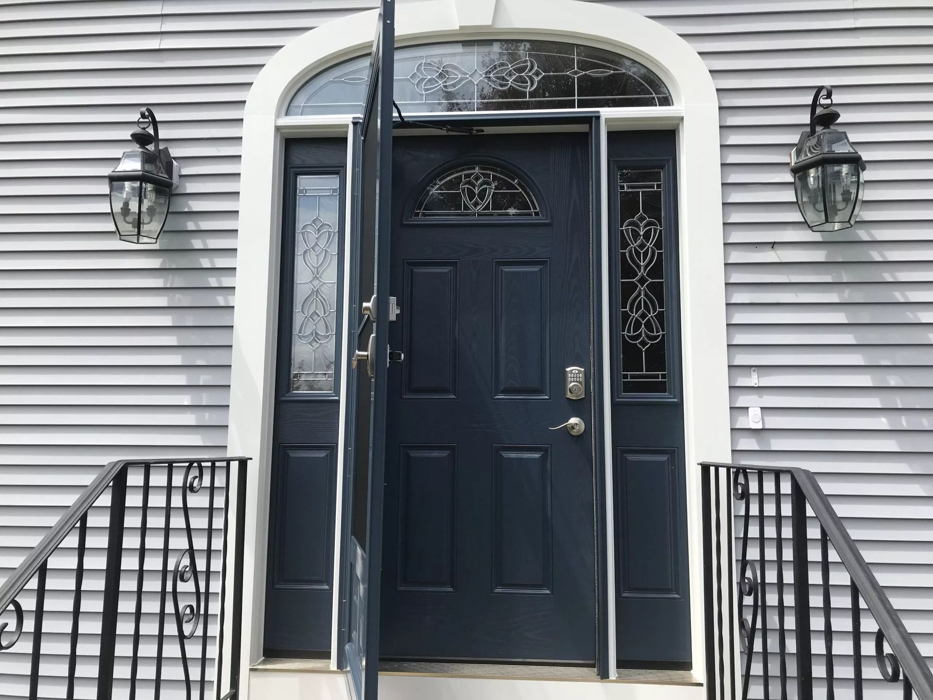 Blue front door with sidelights, porch lights, and wrought iron railing against gray siding. Door is ajar.
