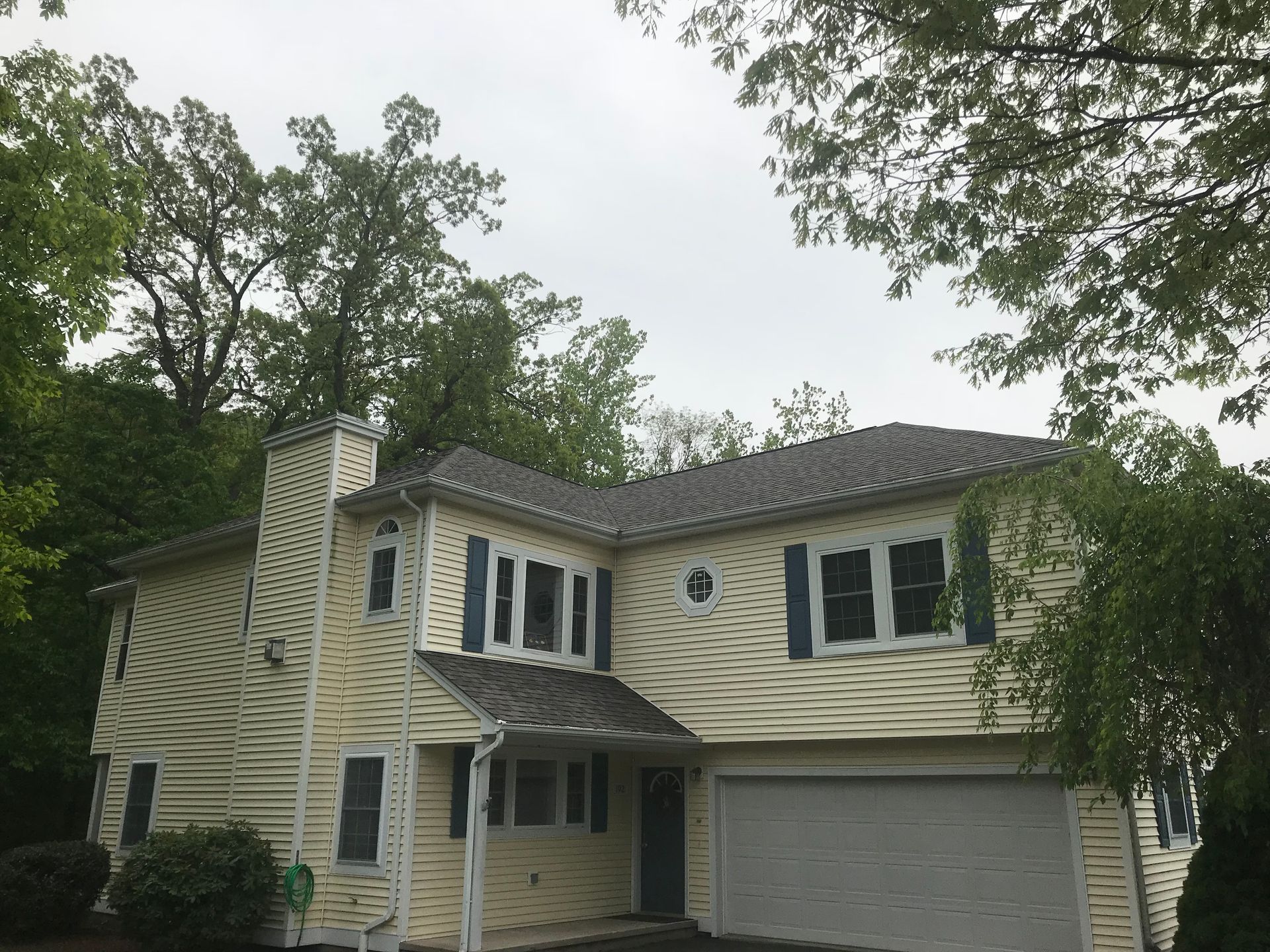 Two-story house with yellow siding, blue shutters, and a gray roof, surrounded by trees under a cloudy sky.