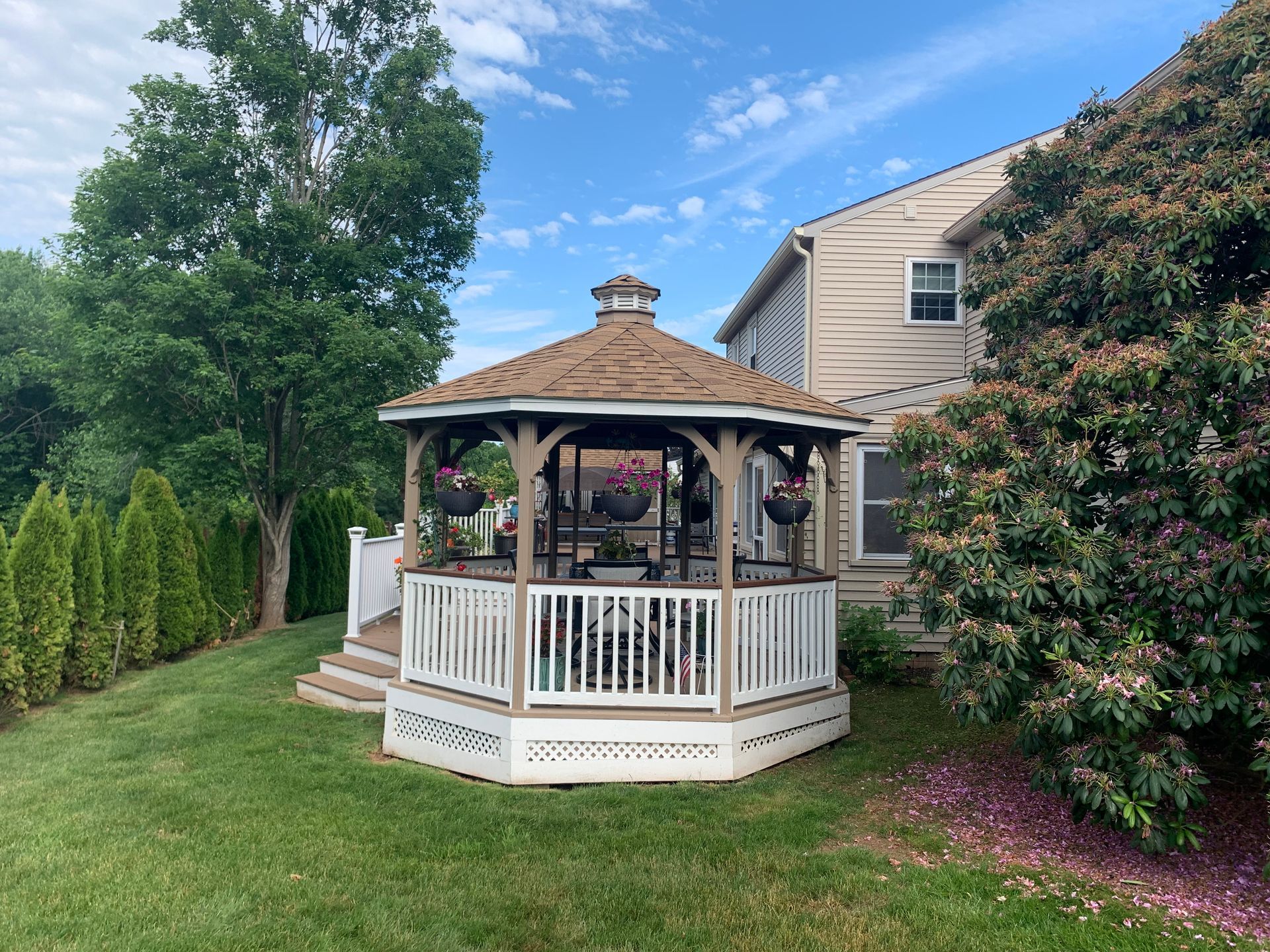Wooden gazebo with brown roof and white railing in a backyard with green grass and trees.