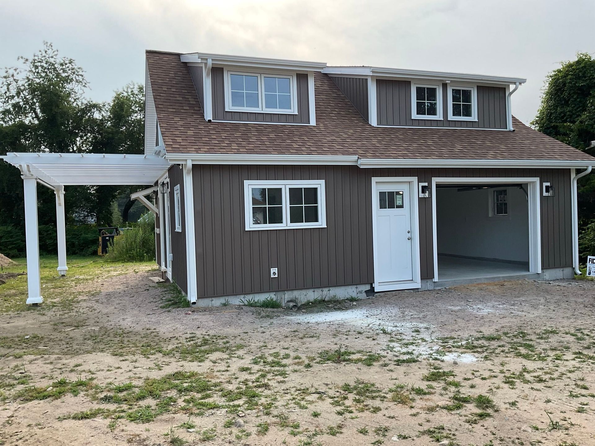 Two-story brown building with garage, white door, and pergola. Gray gravel yard, overcast sky.