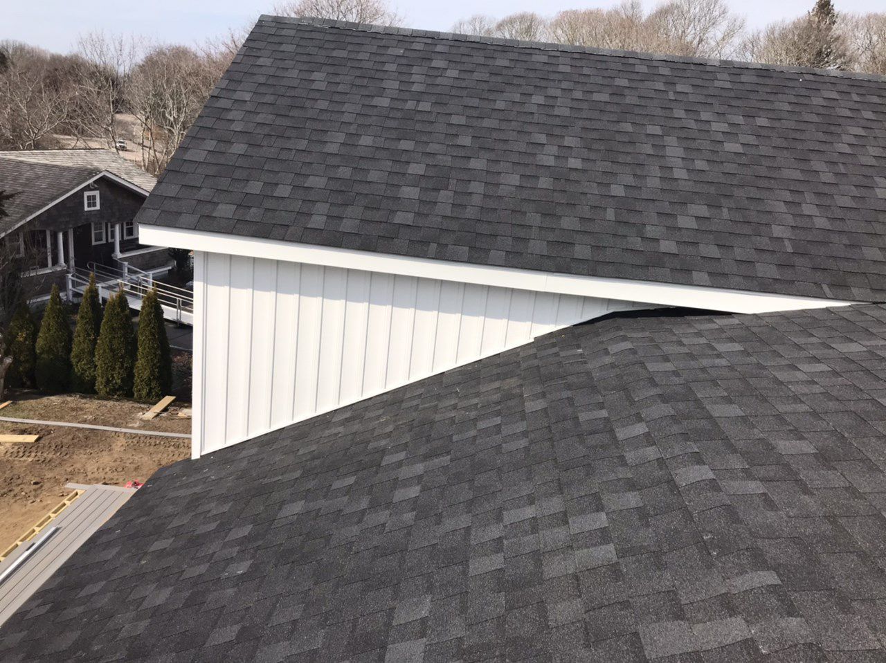 Dark gray shingle roofs meet, with white siding below and a house and trees in the background.