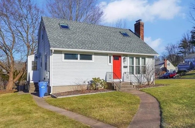 A light gray house with a bright red door, chimney, and blue trash bin. Pathway on green lawn.
