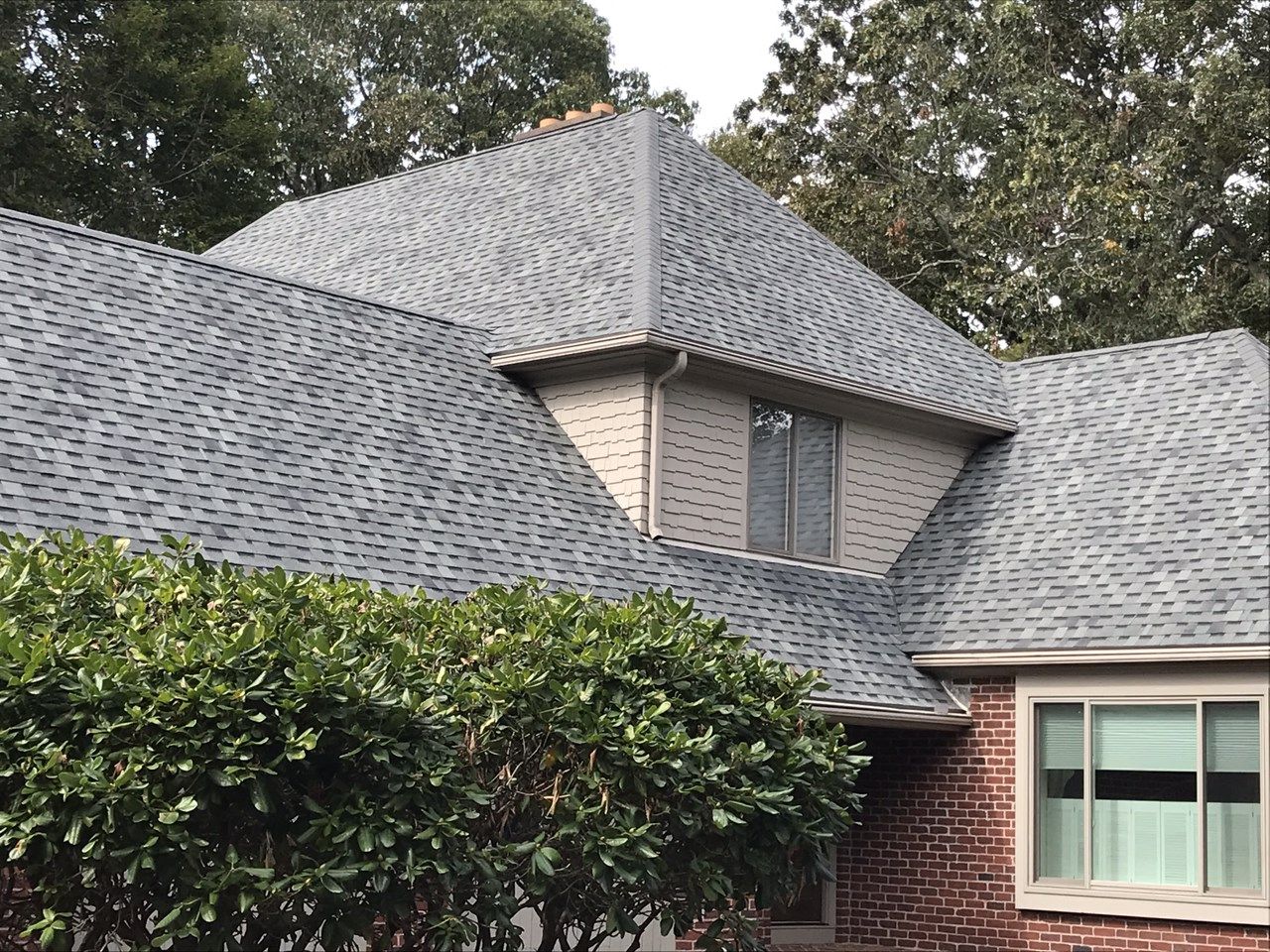 Gray asphalt shingle roof on a house with brick and tan siding, with green bushes in the foreground.