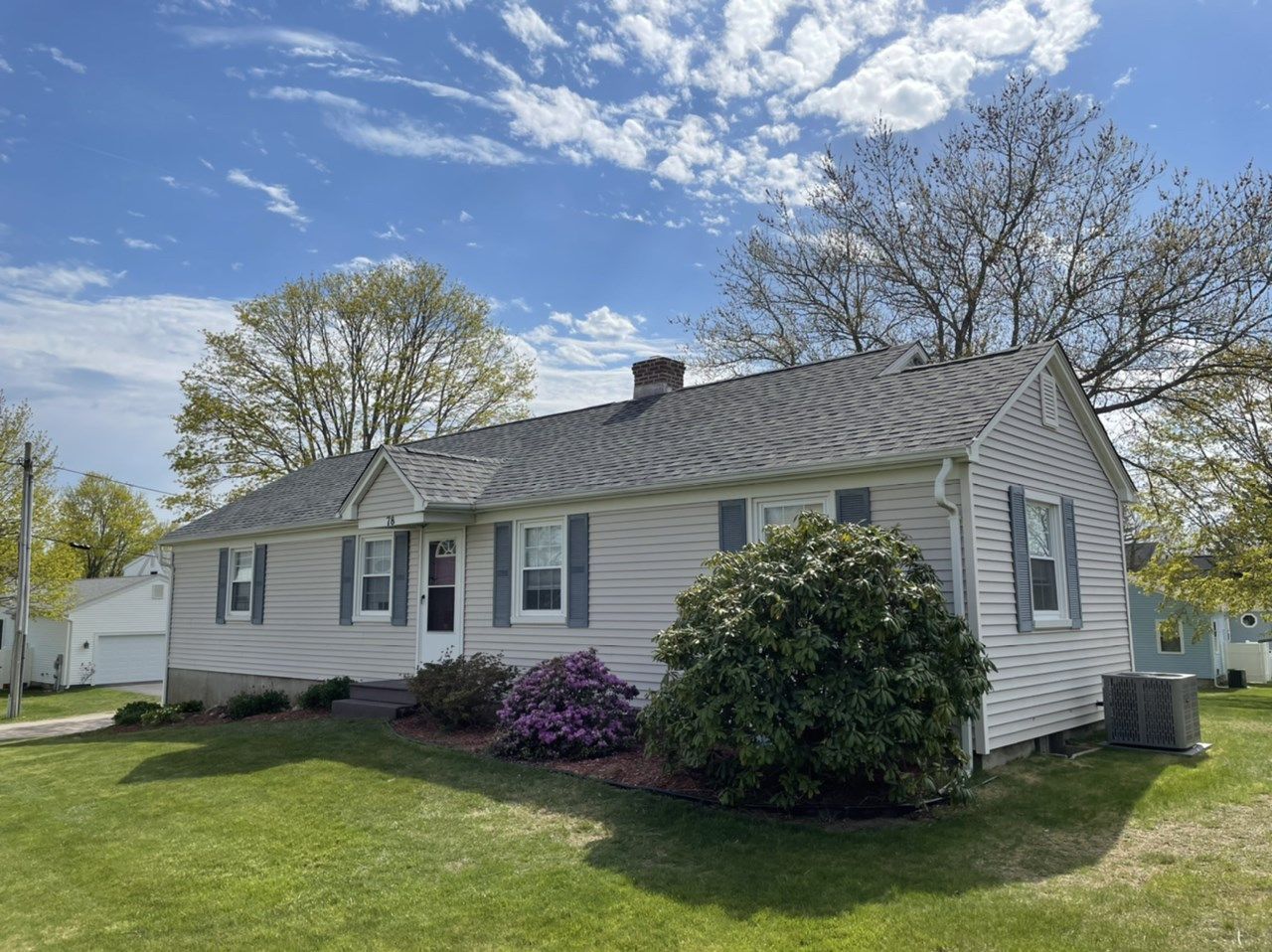 Beige ranch-style house with blue shutters, green lawn, and flowering bushes under a partly cloudy sky.