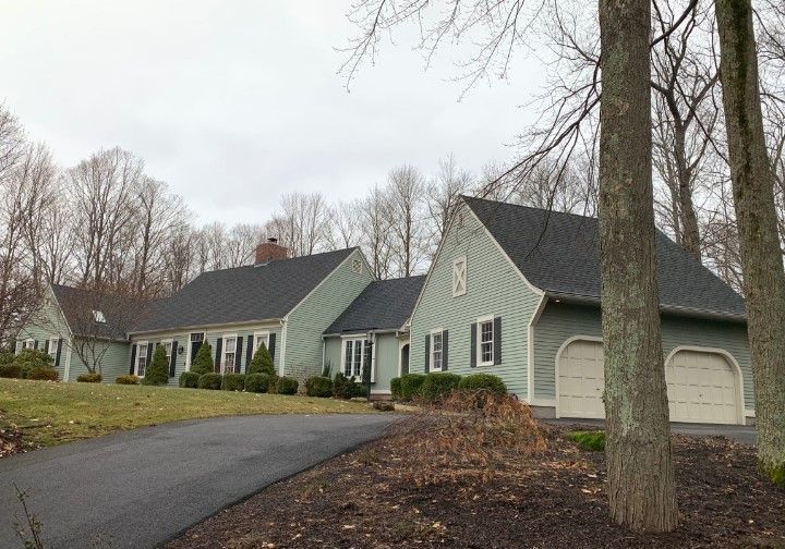 Light green house with black shutters, dark roof, and two-car garage. Driveway leading to the house. Trees in the background.