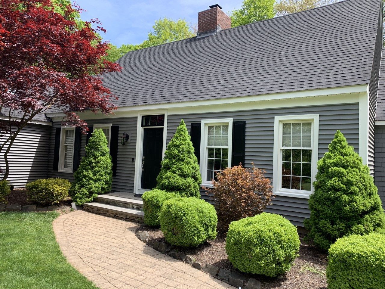Gray house with black shutters, dark roof, brick pathway, and manicured bushes and trees.