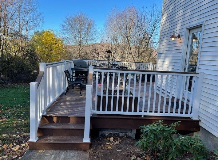 Wooden deck with white railing, steps, grill, and chairs against a house with a door, outdoors.