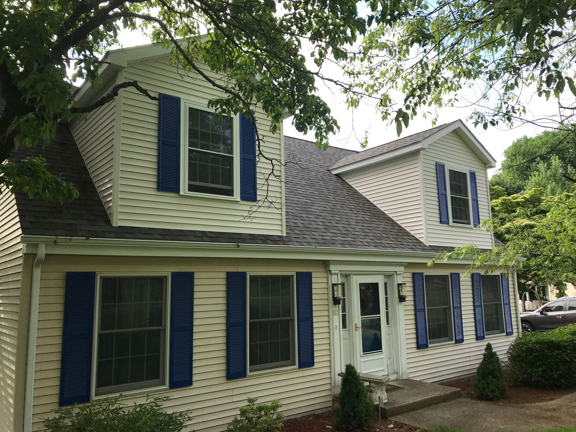Two-story house with cream siding, blue shutters, and dark roof. Trees surround the house.