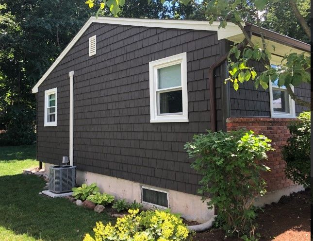 Dark brown shingle siding on a house with white trim. A/C unit, small windows, and landscaping.