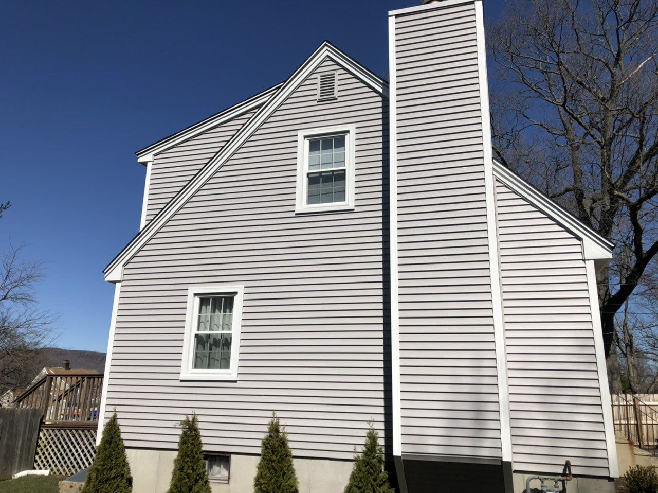 Side view of a two-story gray house with a tall chimney, white trim, and a clear blue sky.