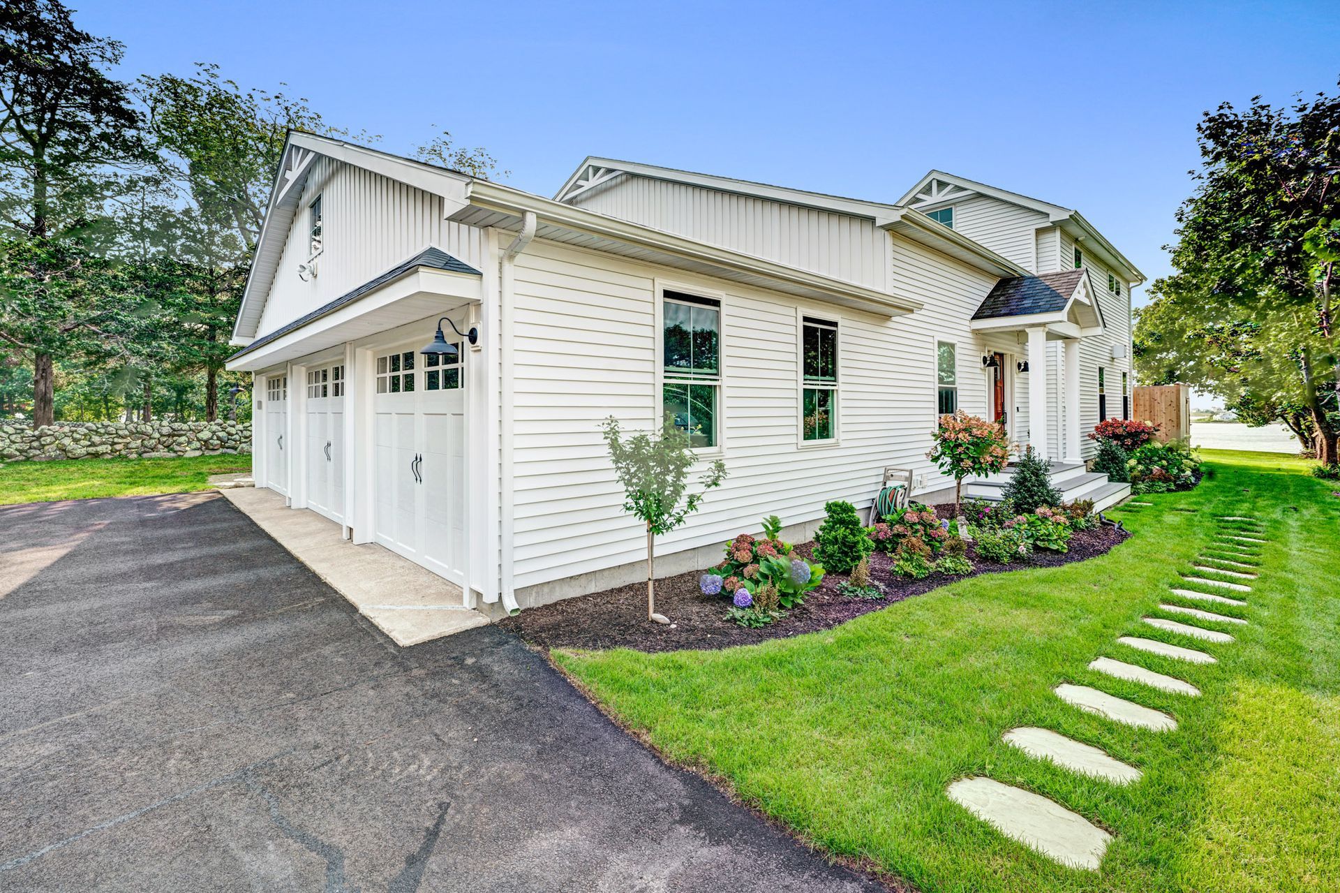 White house with attached garage, landscaped yard, and asphalt driveway under a blue sky.