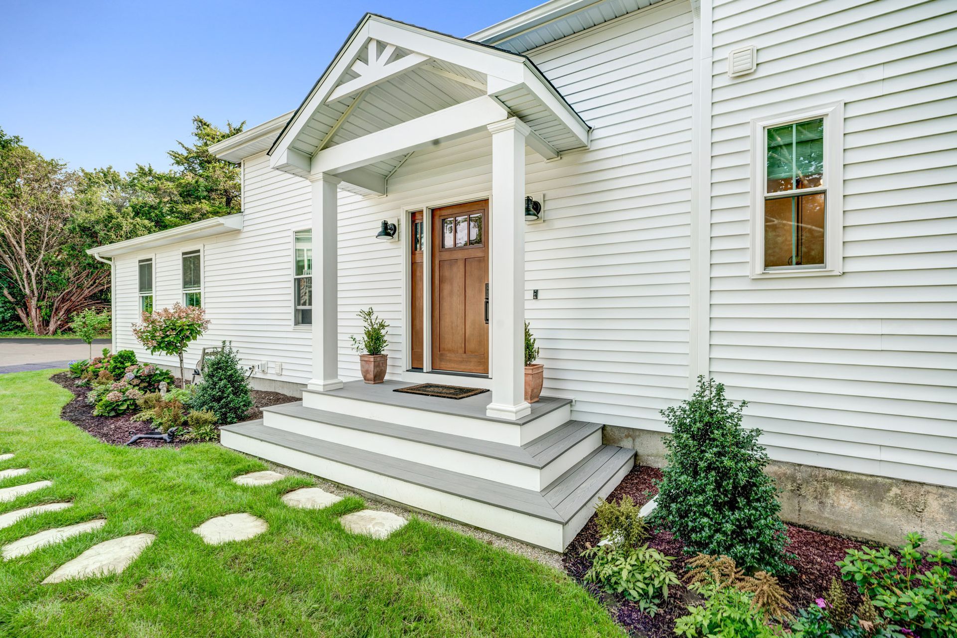White house with a porch and steps. Green lawn with stepping stones. Brown door, white trim.