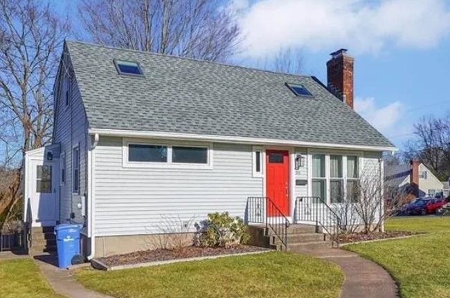 Gray house with red door, chimney, and gray roof; blue trash bin on the side, pathway leading to the front door.
