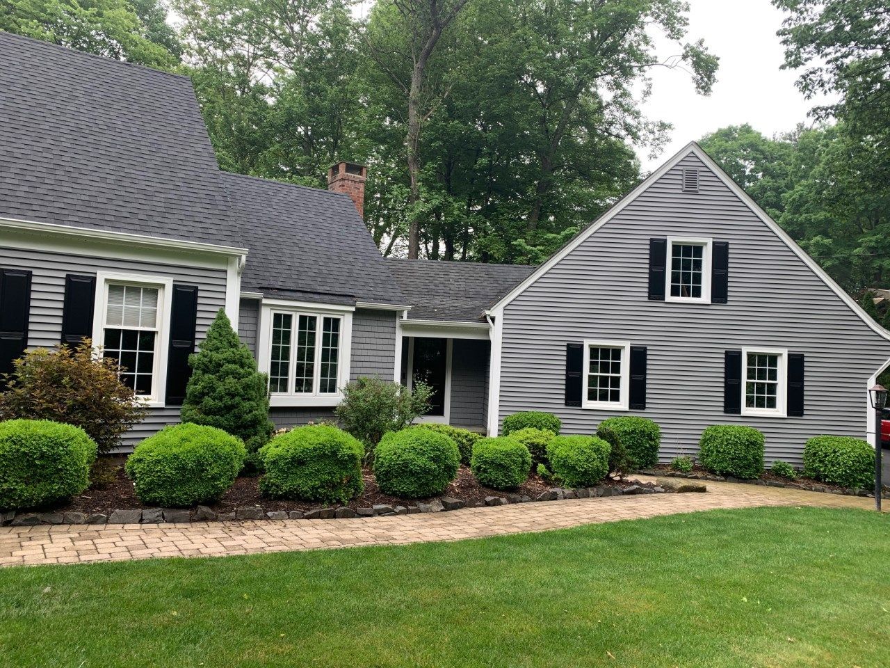 Gray house with black shutters and a brick walkway. Green bushes line the path.