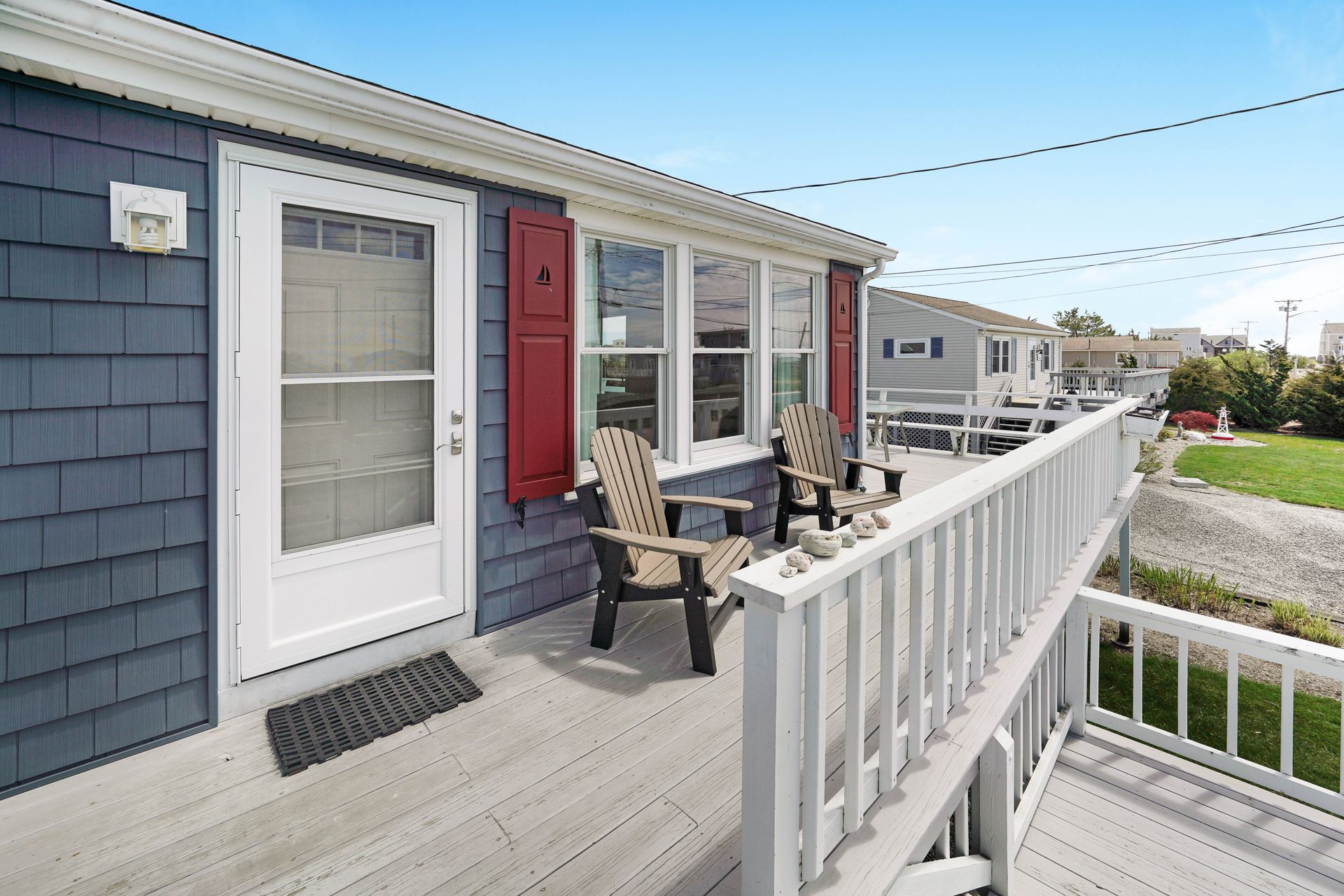 Blue-shingled beach house with a white porch, door, and railing, and two chairs. Red shutters.