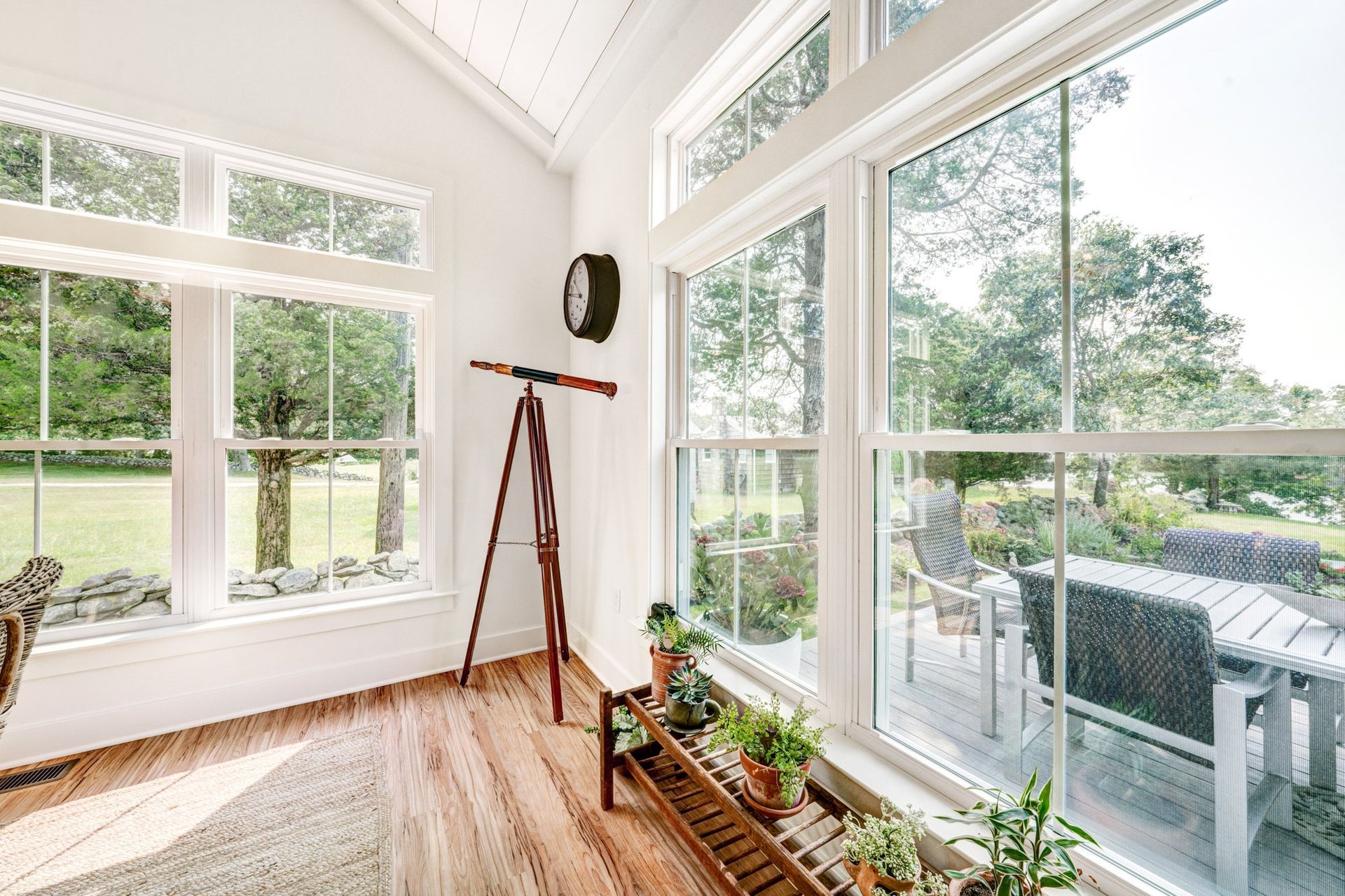 Bright sunroom with large windows overlooking a backyard; a telescope stands by the window with some potted plants on a small shelf.
